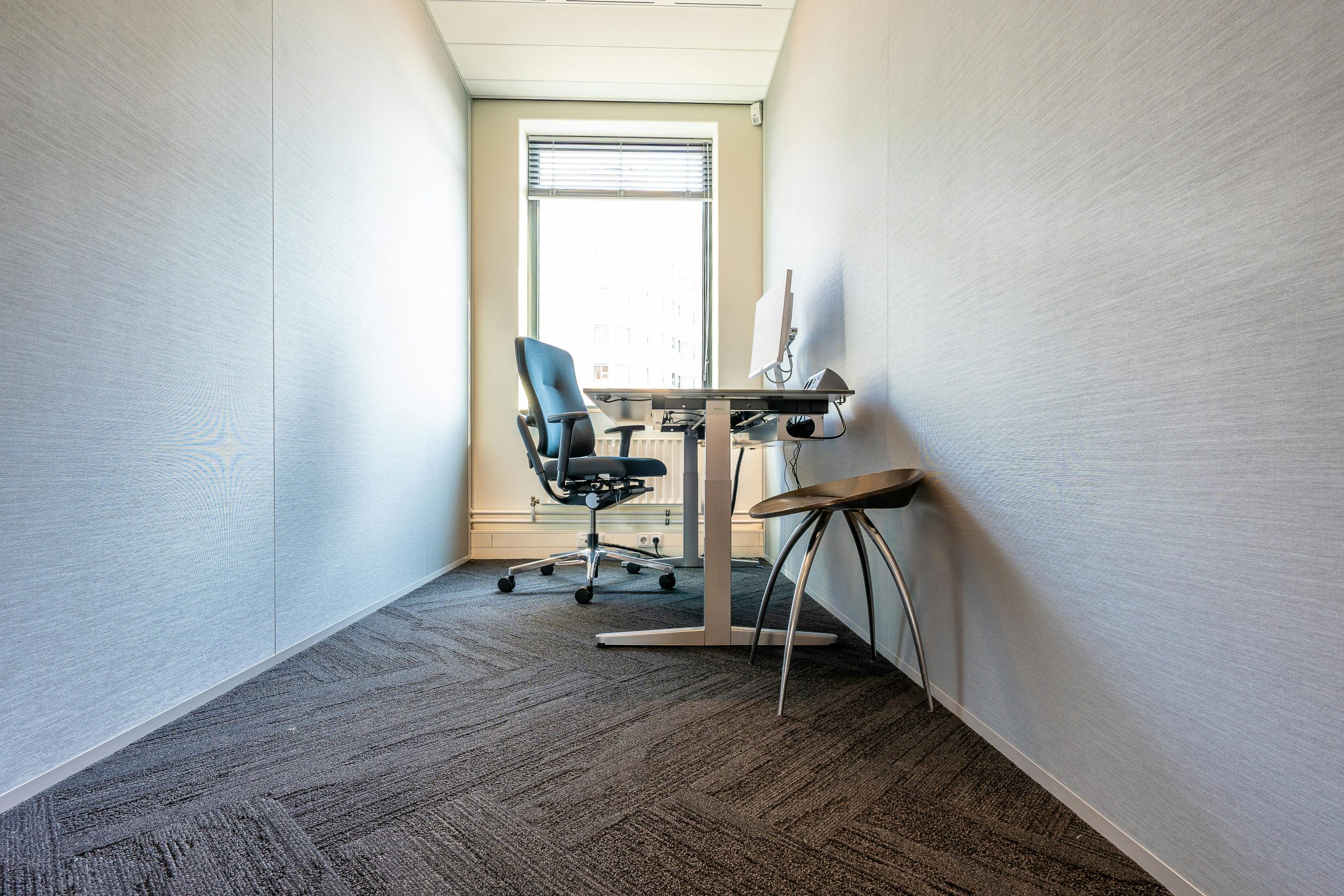 A clean, minimalist office space featuring a desk, chair, and computer by a large window.