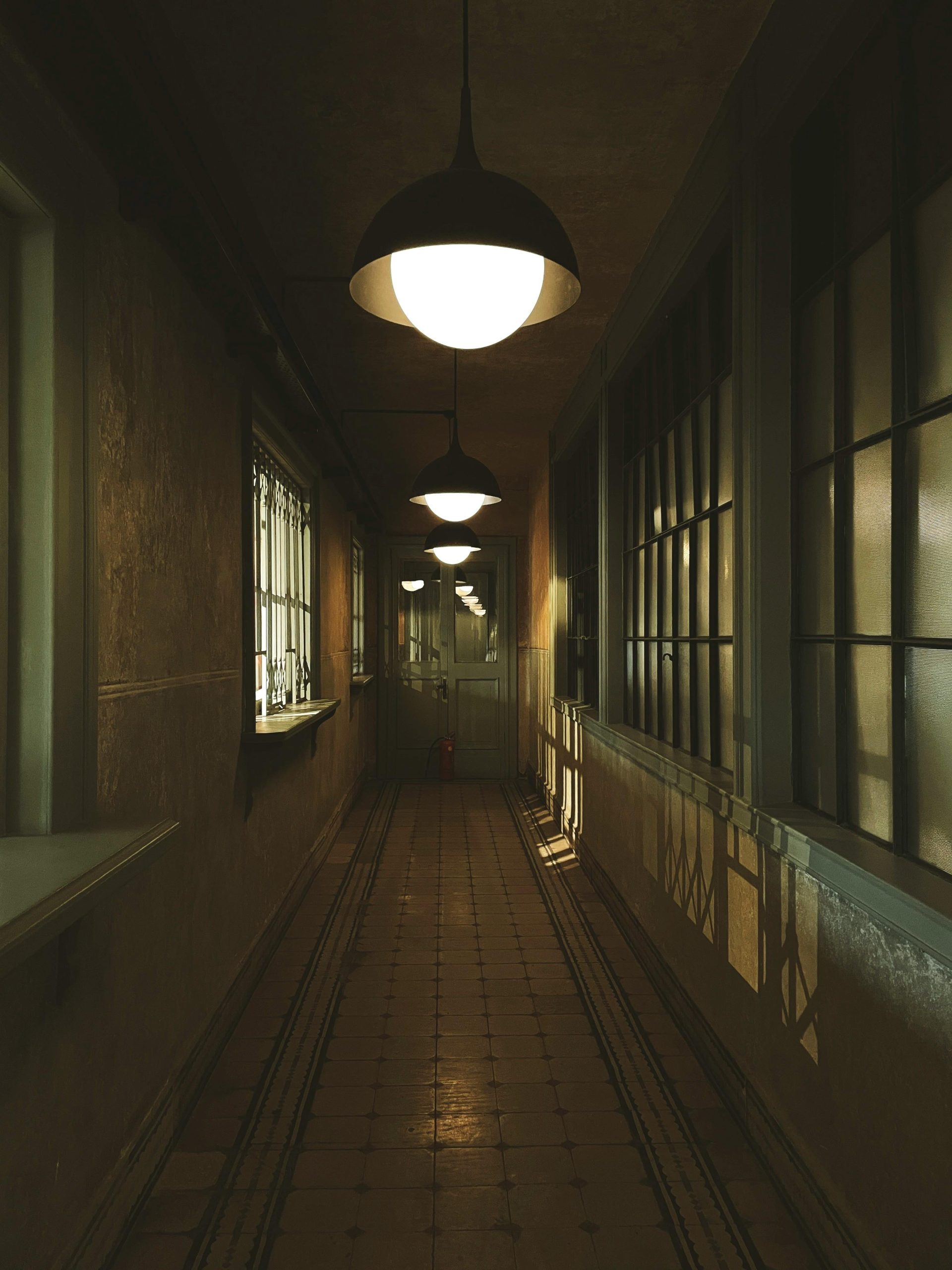 A narrow, dimly lit hallway with vintage pendant lights and large windows.