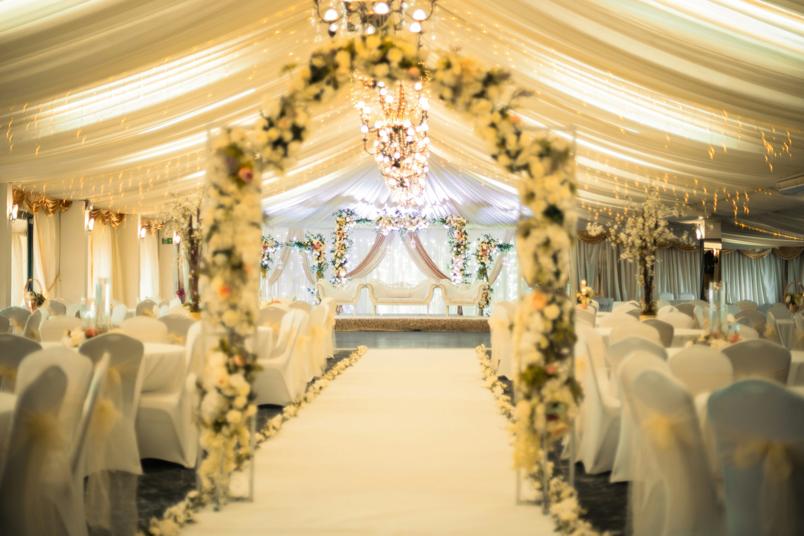 Breathtaking wedding ceremony hall at Goosedale, Nottinghamshire featuring a white aisle flanked by lush floral arches of cream and blush roses, draped ivory ceiling with cascading fairy lights, cryst
