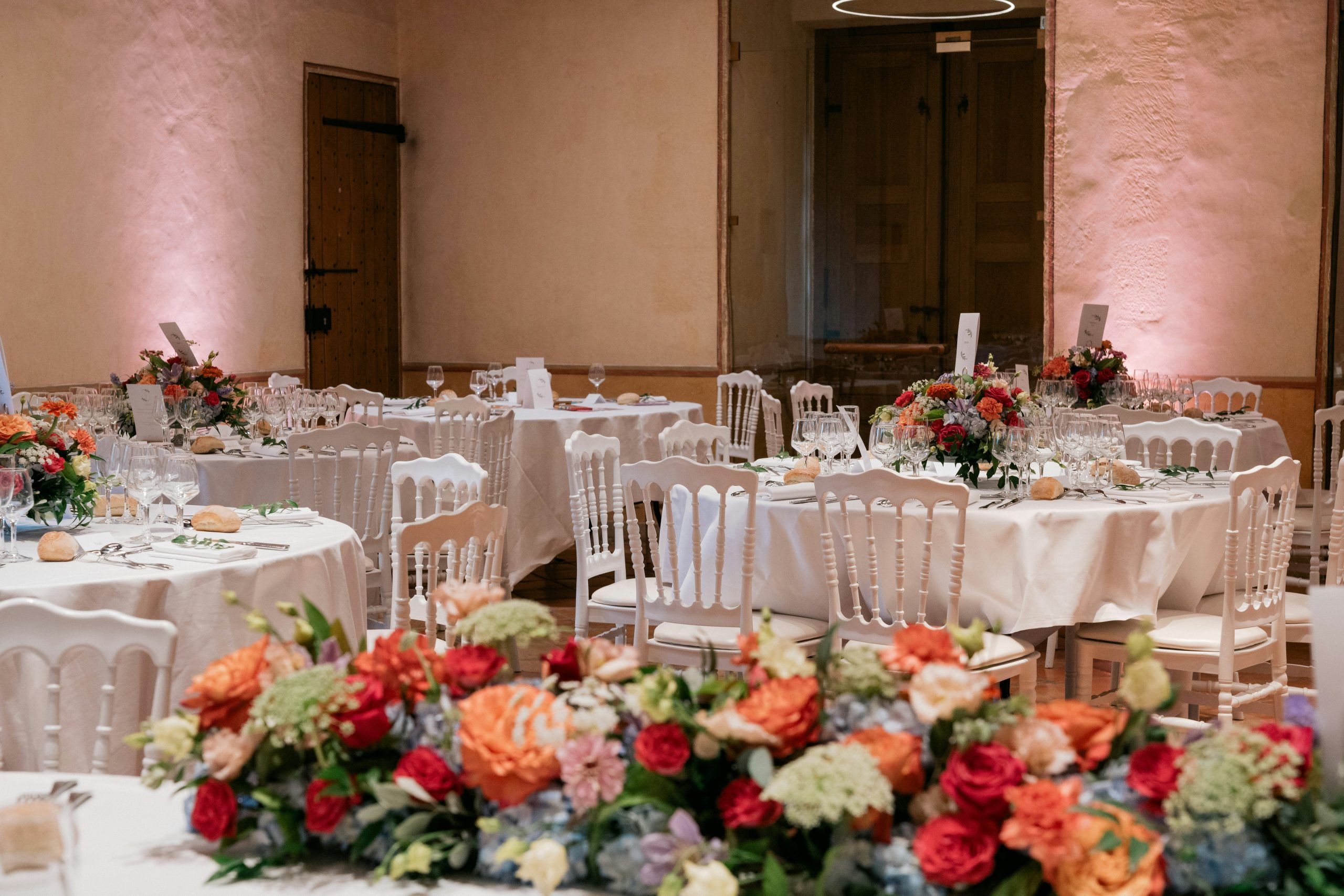 ChatGPT a dit : C’est une salle de réception élégante, décorée pour un mariage, avec des tables rondes dressées de blanc, des chaises classiques et de grands centres de table floraux aux couleurs vive