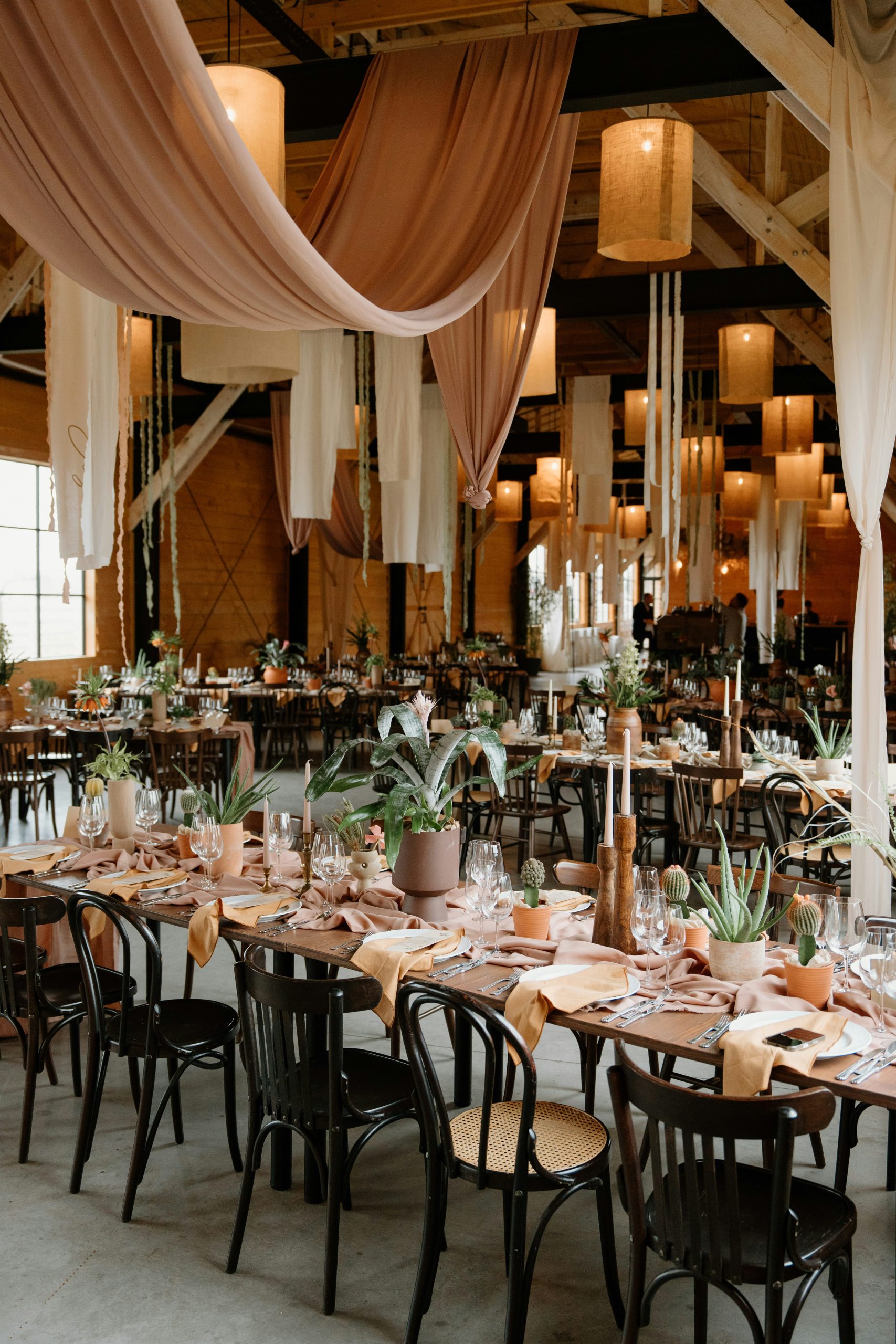 Elegant rustic dinner setup inside a modern wooden barn, styled for a western-inspired event. Long wooden tables are carefully arranged with neutral-toned tableware, linen napkins, candles, and lush p