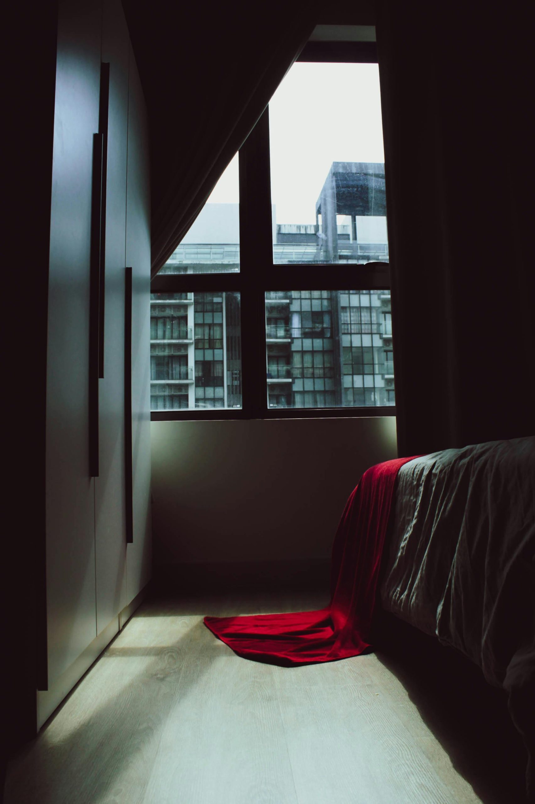 Explore a minimalist bedroom with a dark ambiance and a striking red blanket near a large window overlooking city buildings.