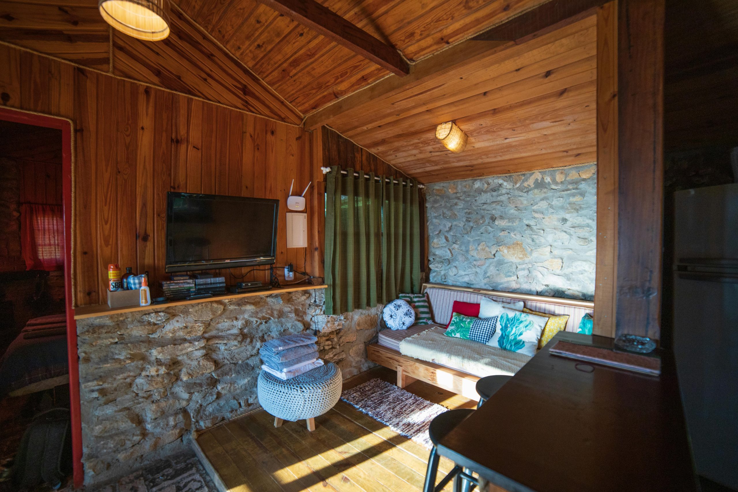 Sofa with pillows placed in room near TV and counter in rustic style house with wooden walls decorated with stones