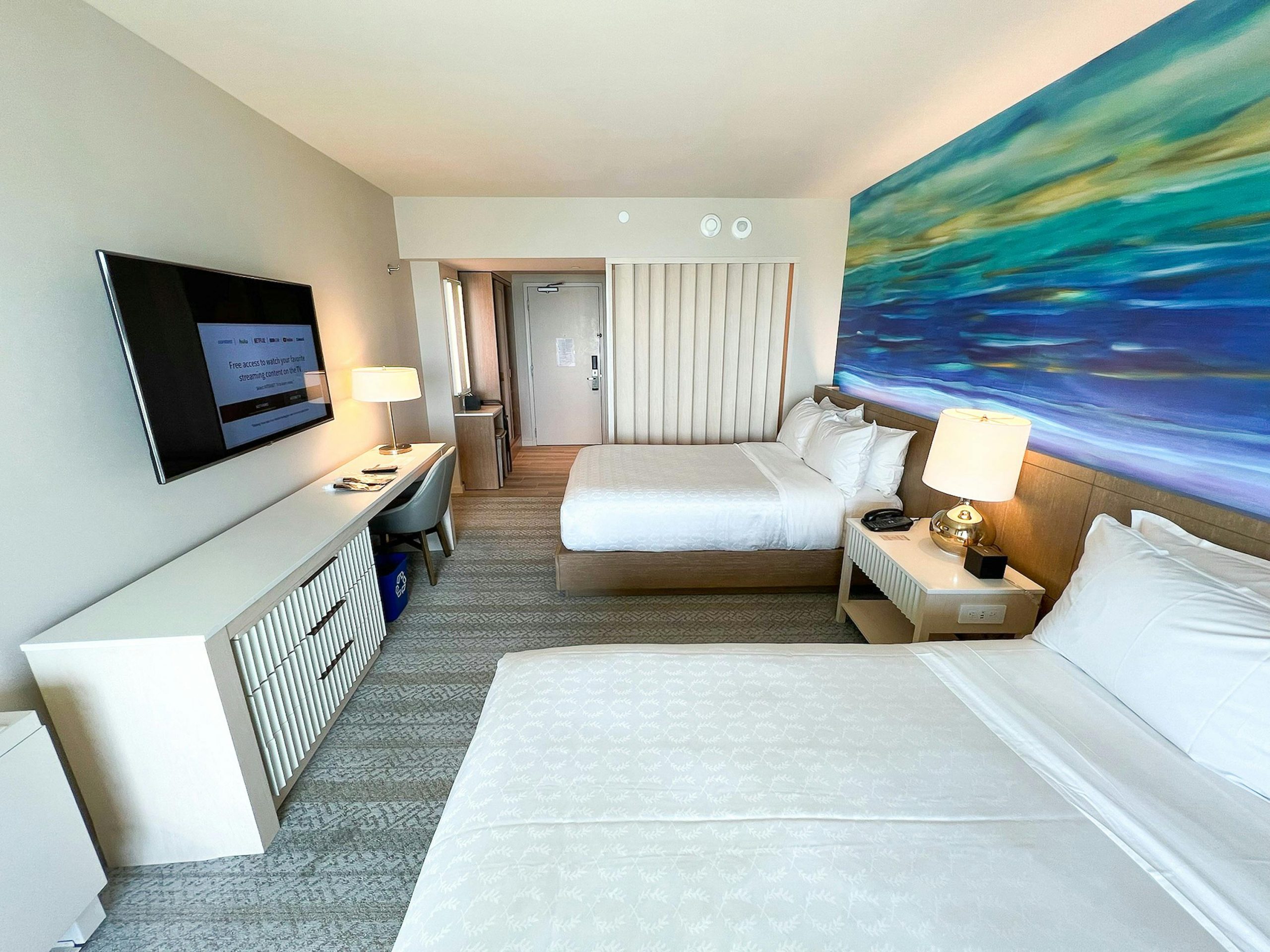 Spacious hotel room in Honolulu featuring two beds, vibrant ocean artwork, and modern amenities.