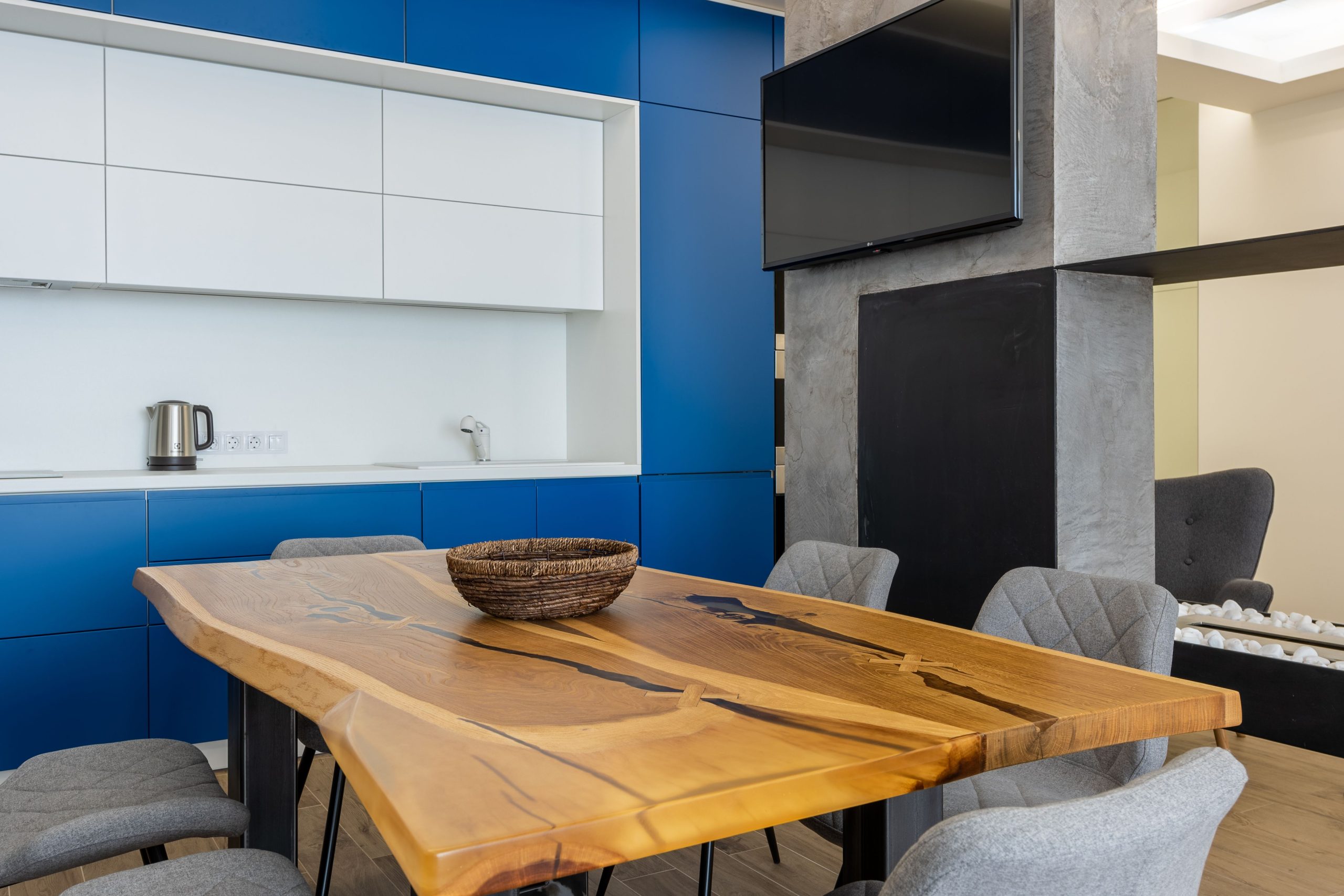 Wooden table with bowl and chairs placed near blue and white cupboards with kettle on counter in contemporary kitchen with TV