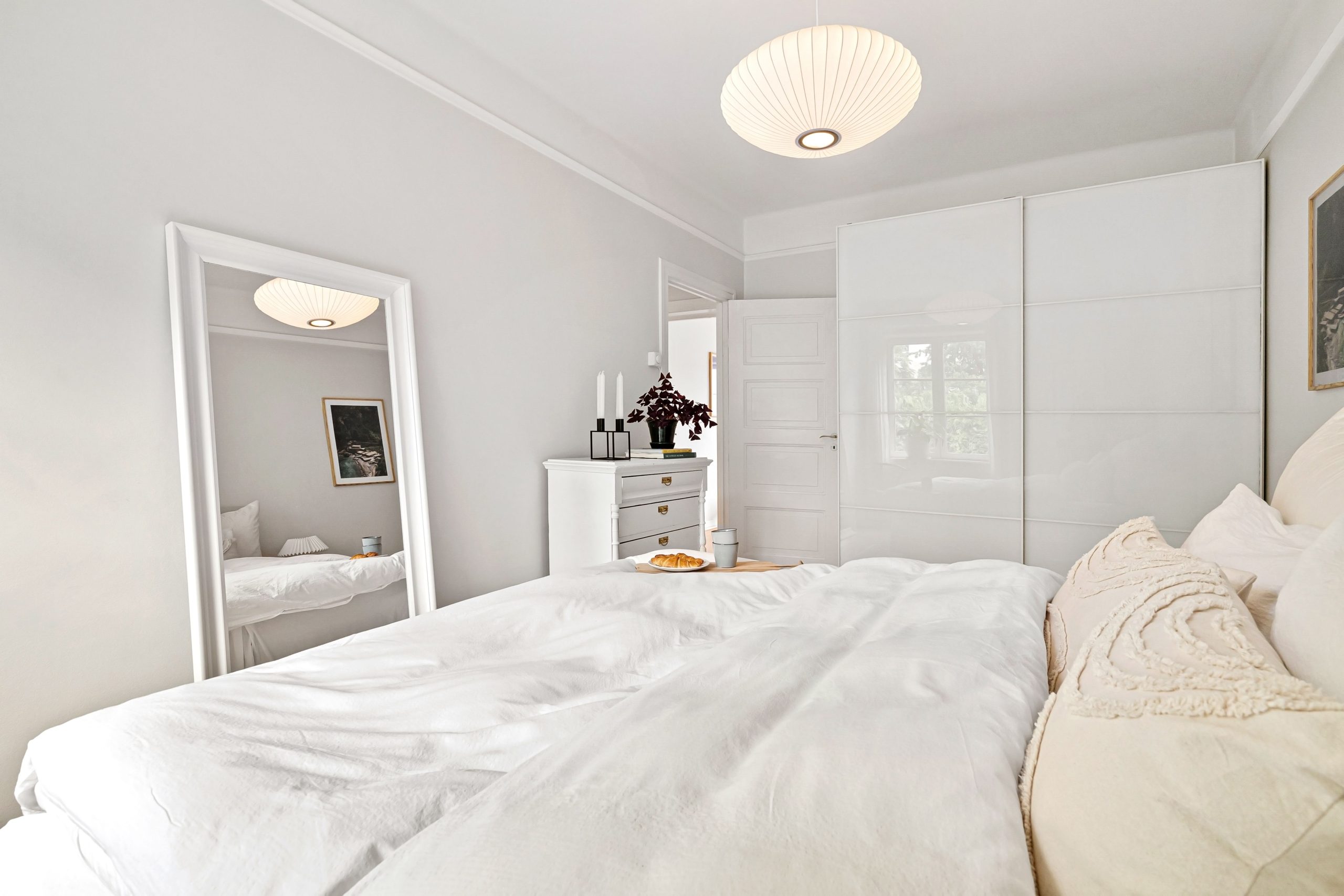 A minimalist bedroom with white decor, featuring a bed, dresser, and mirror under soft lighting.