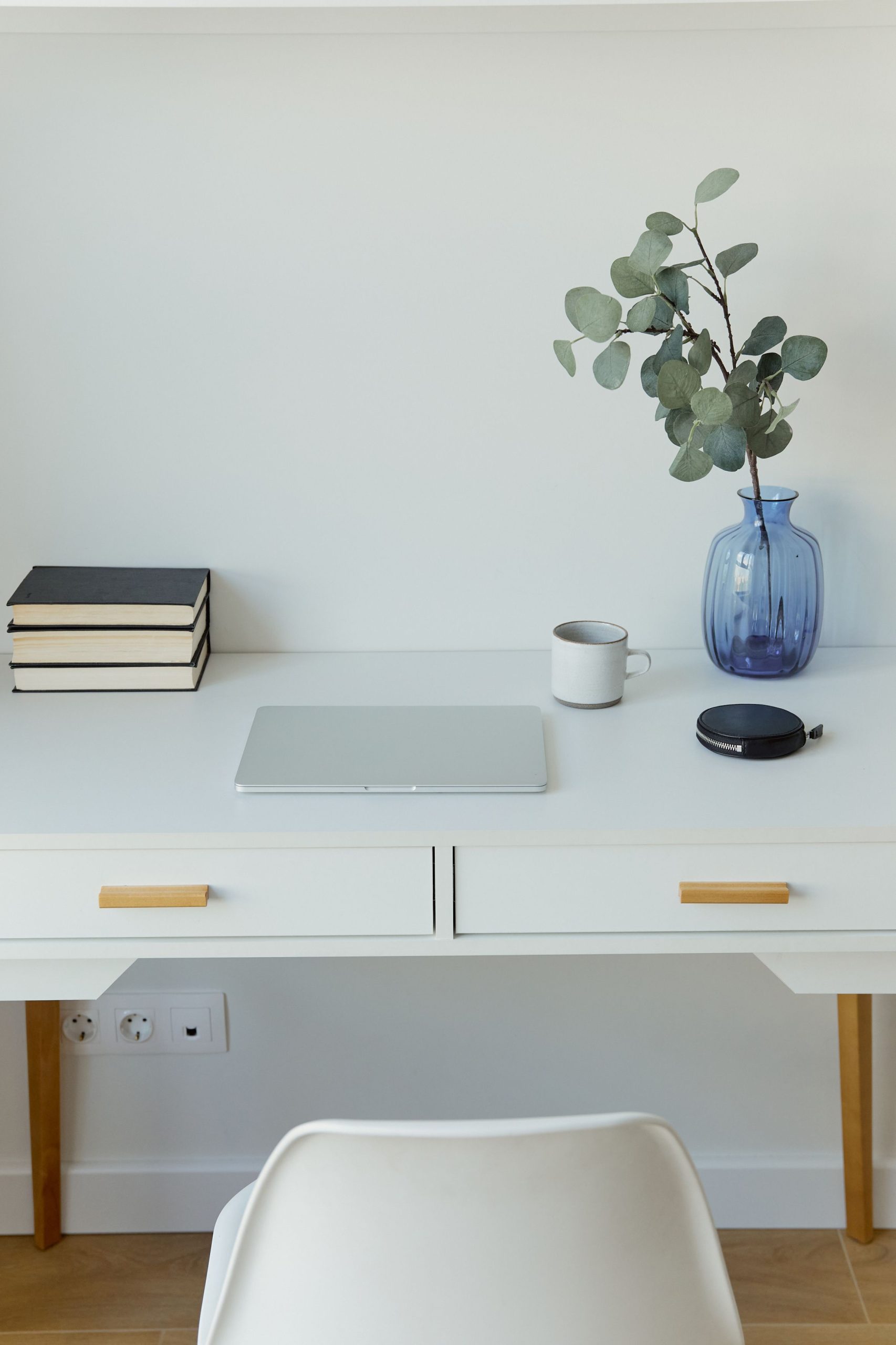 Clean workspace featuring a white desk, laptop, books, mug, and a vase with greenery.