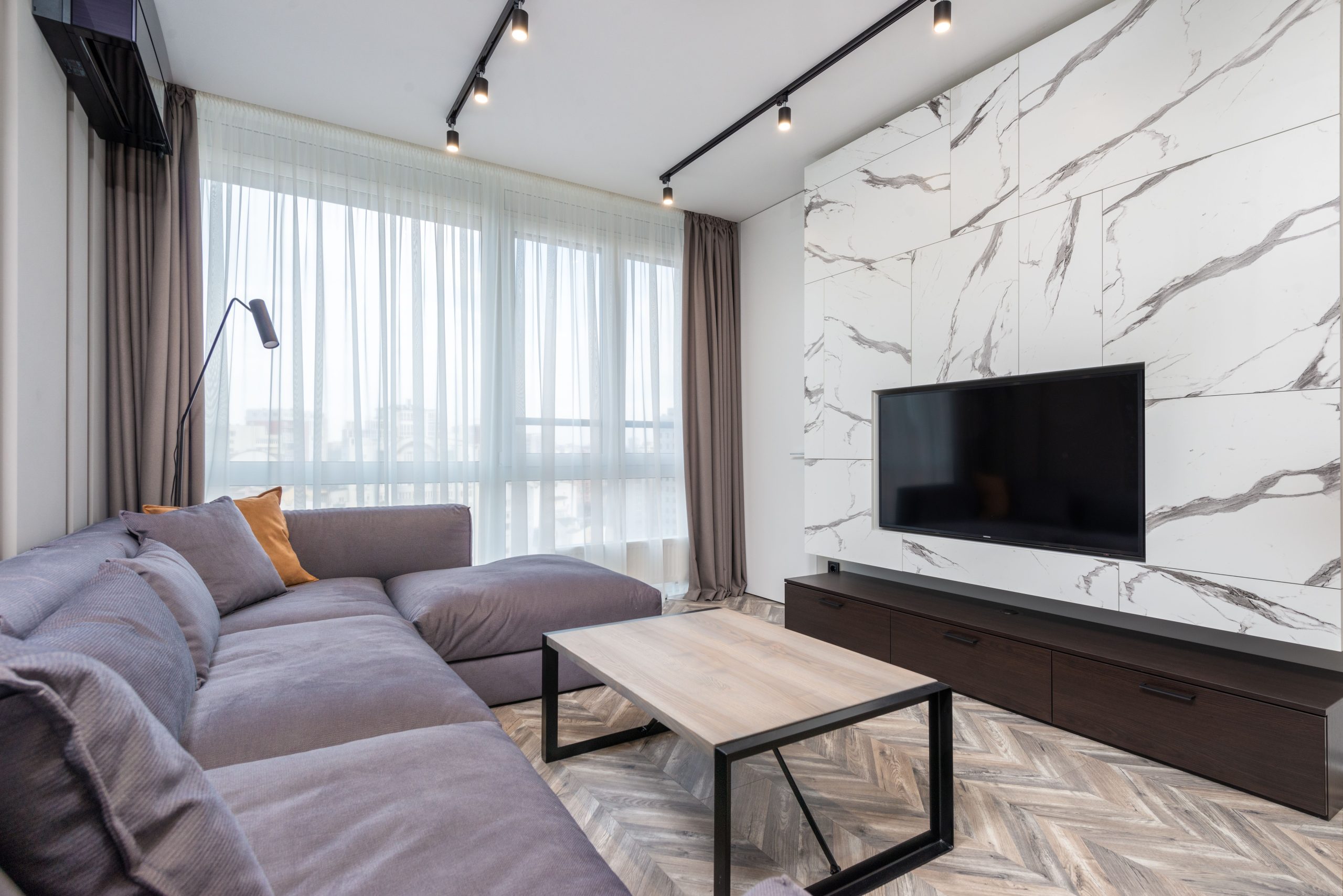 Contemporary living room featuring a marble wall with a large TV, cozy sectional sofa, and ample natural light.
