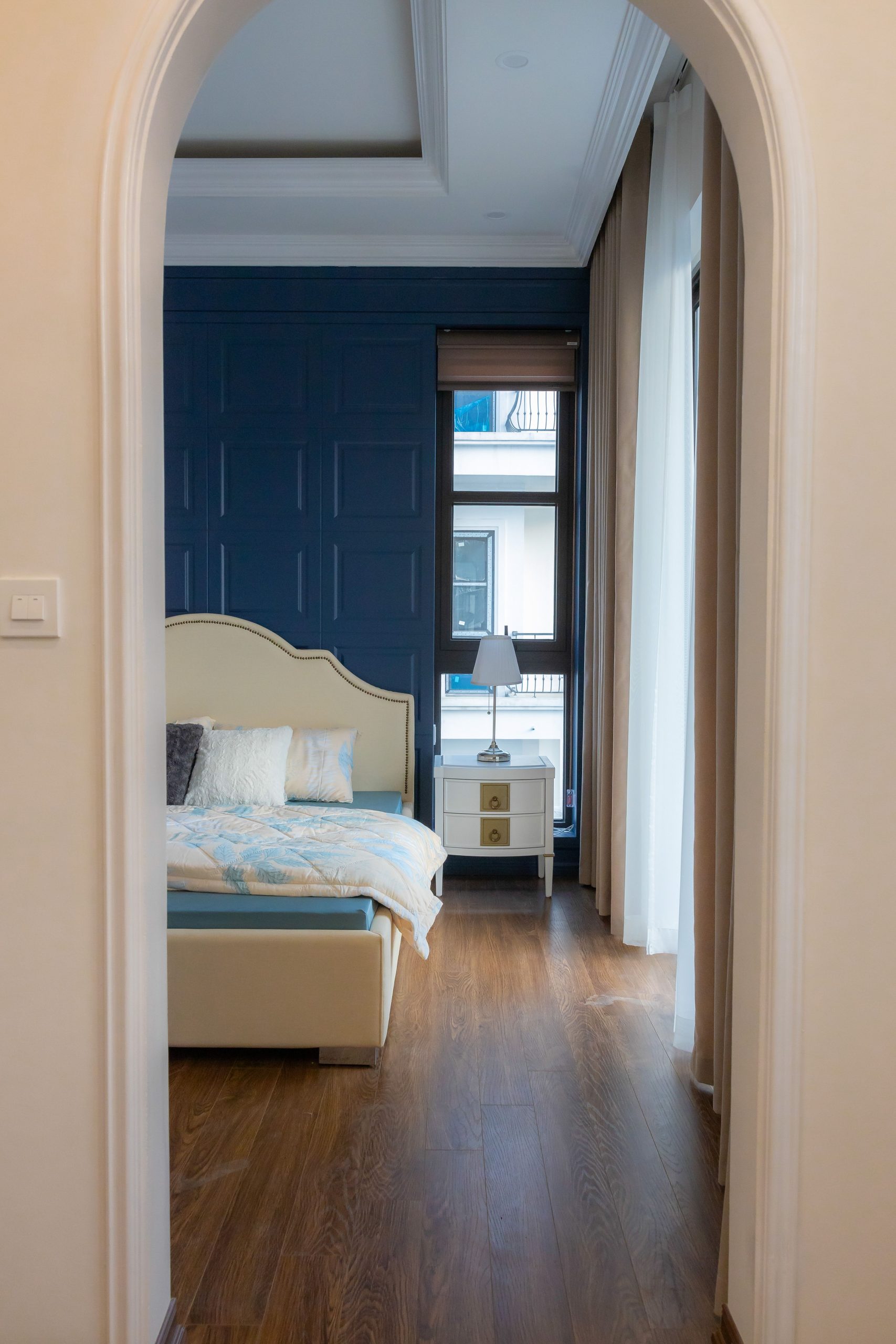 Elegant bedroom with blue accent wall and wooden flooring, viewed through a white archway.