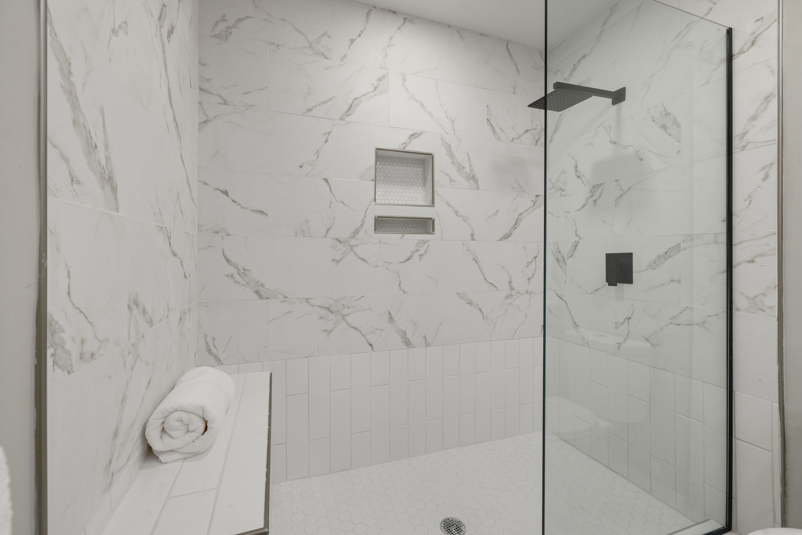 Elegant white marble shower with glass door, bench, and towel in modern bathroom.