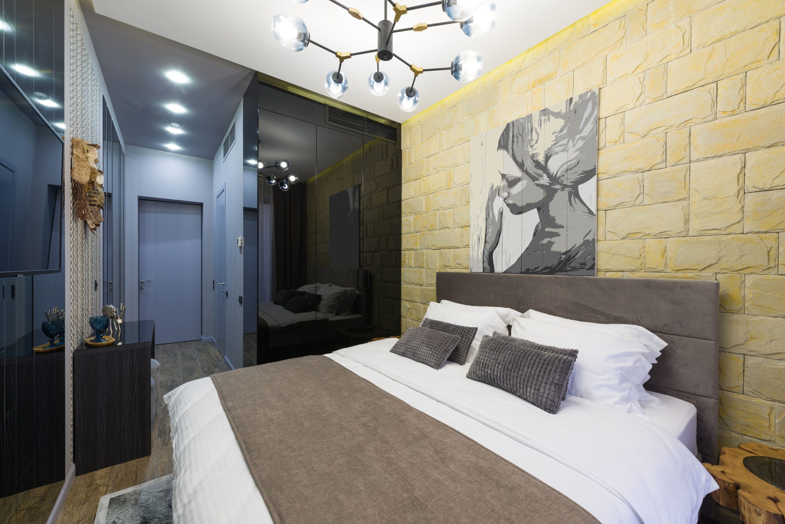 Loft style room interior with bed reflecting on wall against TV set and artwork illuminated by chandelier in hotel