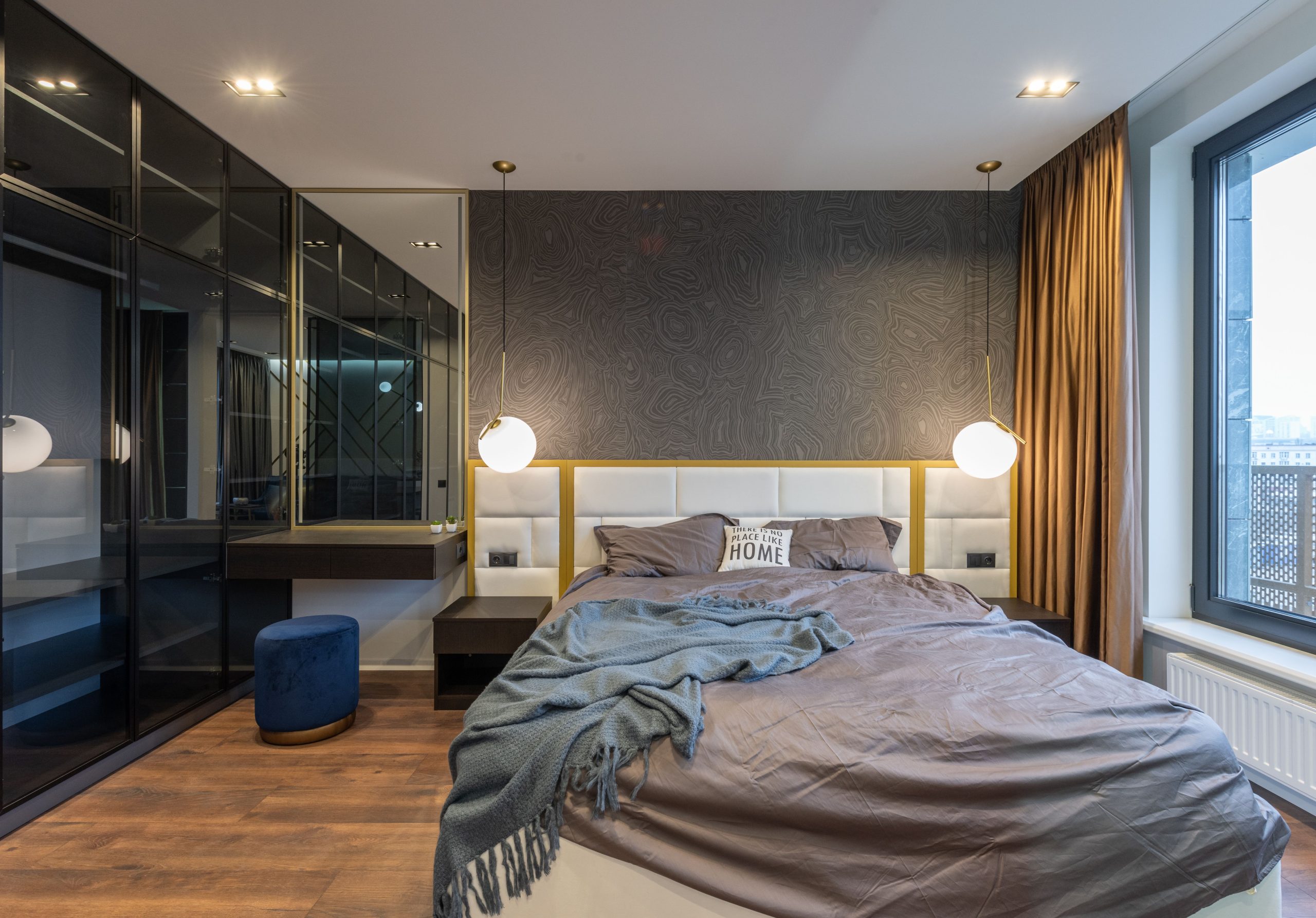 Modern light bedroom interior with soft bed with pillows and blanket near lamps and mirror with shelf near pouf and window with curtains